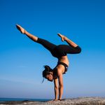 A woman performing a yoga handstand on a rock by the sea under a clear blue sky.